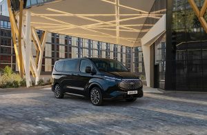 Schwarzer Ford E-Tourneo Custom vor einem modernen, neuen Gebäude. Das Fahrzeug kombiniert beeindruckende Leistungsfähigkeit, ein elegantes Design und hochwertige Ausstattungsdetails und hebt sich als fortschrittliches Elektrofahrzeug im Bereich der Mobilität und des Komforts hervor.