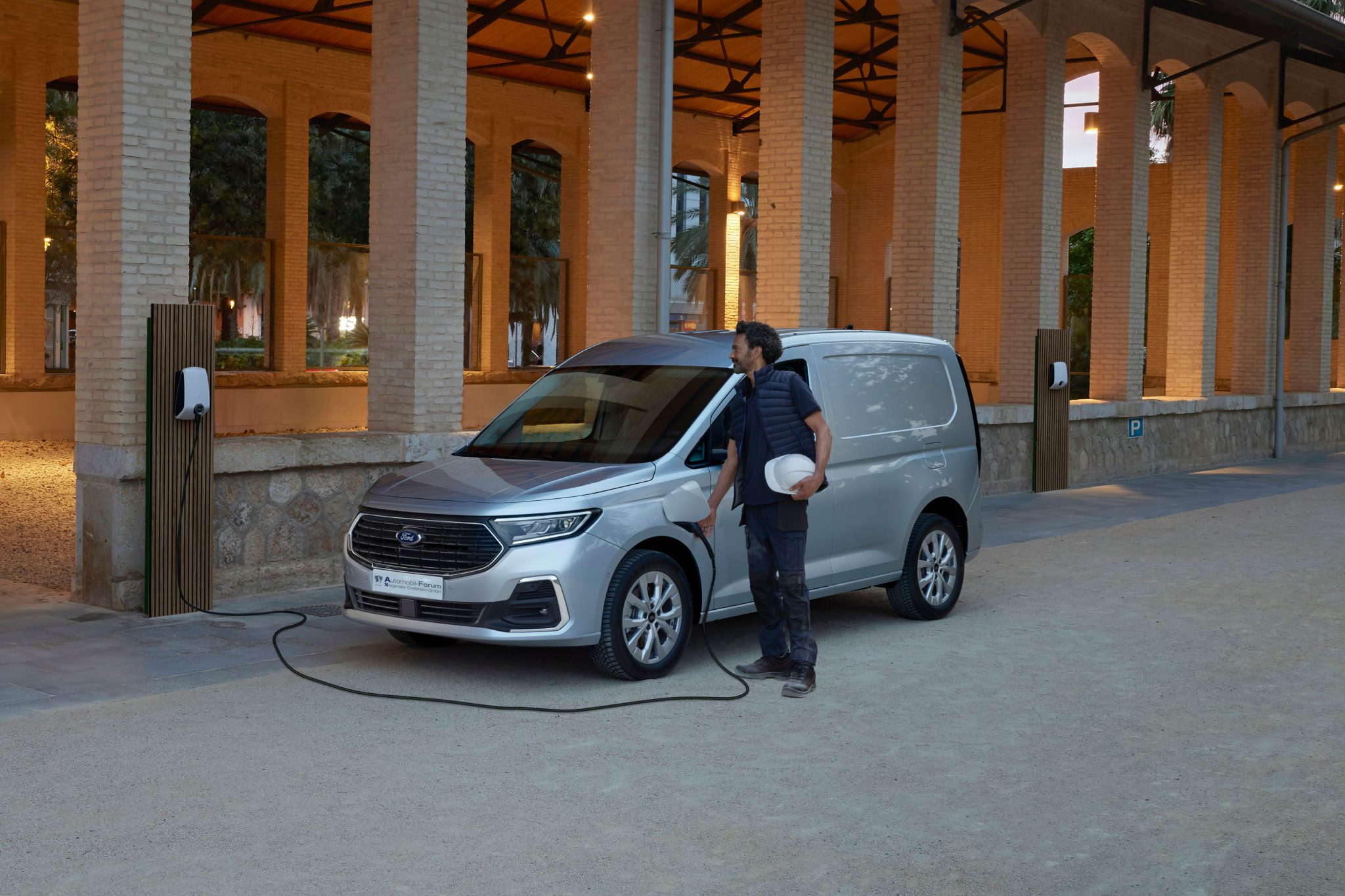 Der neue Ford Transit Connect mit Plug-in-Hybrid steht an einer Ladestation und wird gerade aufgeladen. Das Auto parkt neben einem modernen Gebäude, das zur gewerblichen Nutzung passt. Das Design des Fahrzeugs ist praktisch und funktional, mit klaren Linien und einer Ladestation, die an der Seite angeschlossen ist.