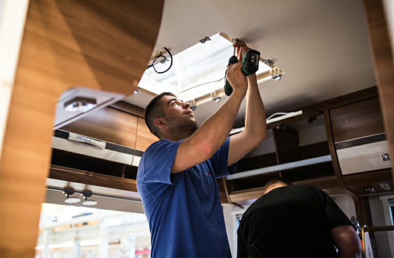 Zwei Handwerker montieren ein Schiebedach an einem Ford Transit Camper-Van – professioneller Ausbau mit Werkzeugen im Handwerks-Camping-Projekt.